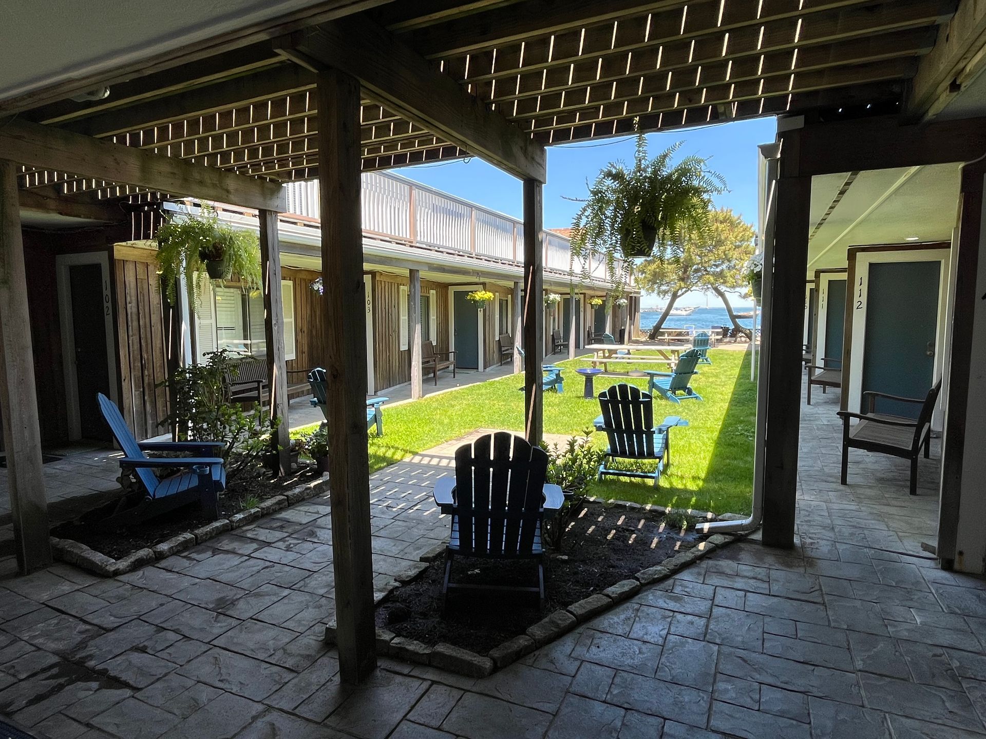 View of a courtyard with blue Adirondack chairs, grass, and a waterfront. Wooden beams provide shade over brick pathways and walkways.