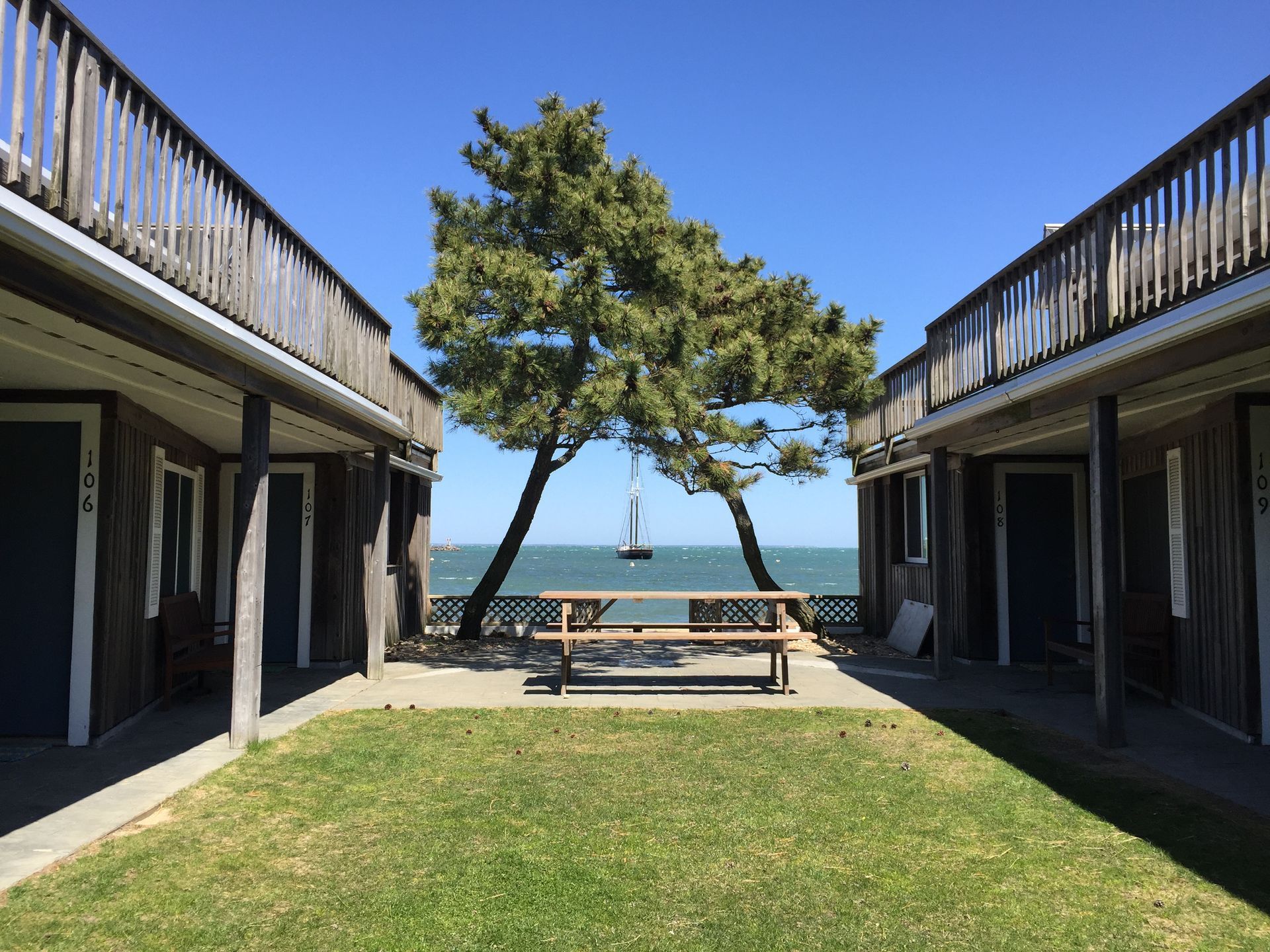 View between two wooden motel buildings toward a sailboat on the water under a tree and picnic table on a sunny day.