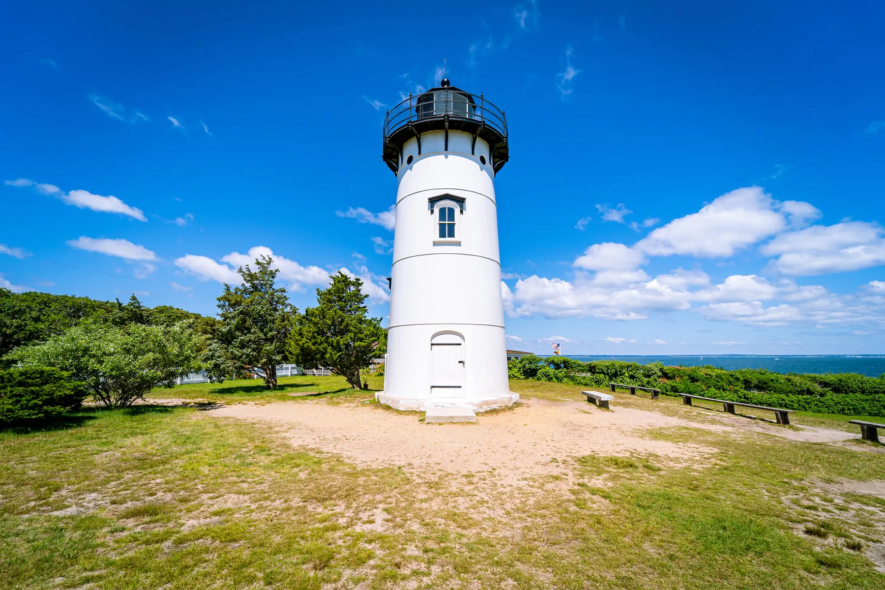 White lighthouse on a grassy hilltop under a bright blue sky. Small trees and ocean visible in the distance.