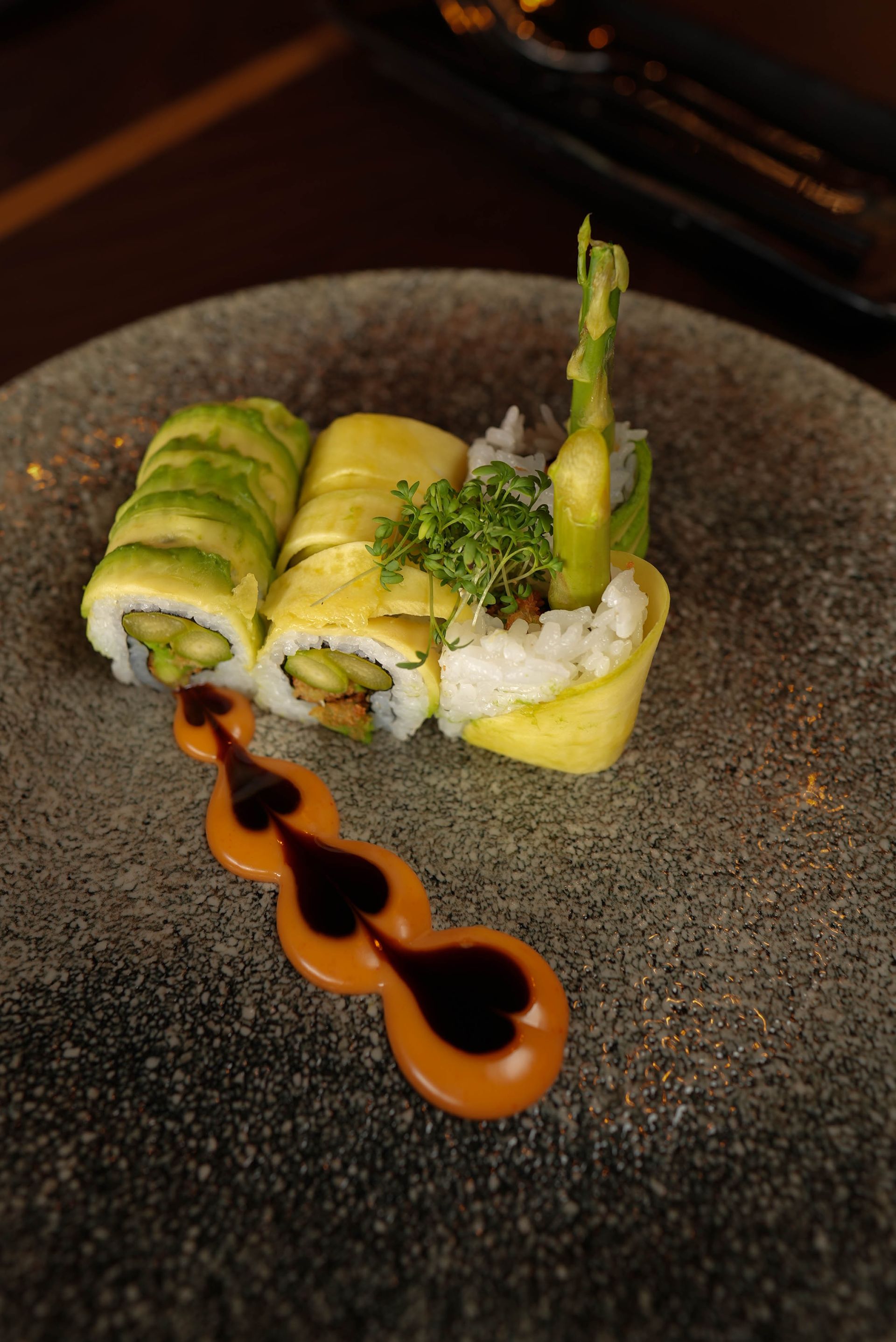 Sushi-Rollen auf einem dunklen Teller mit Spargel und beträufelter Sauce.
