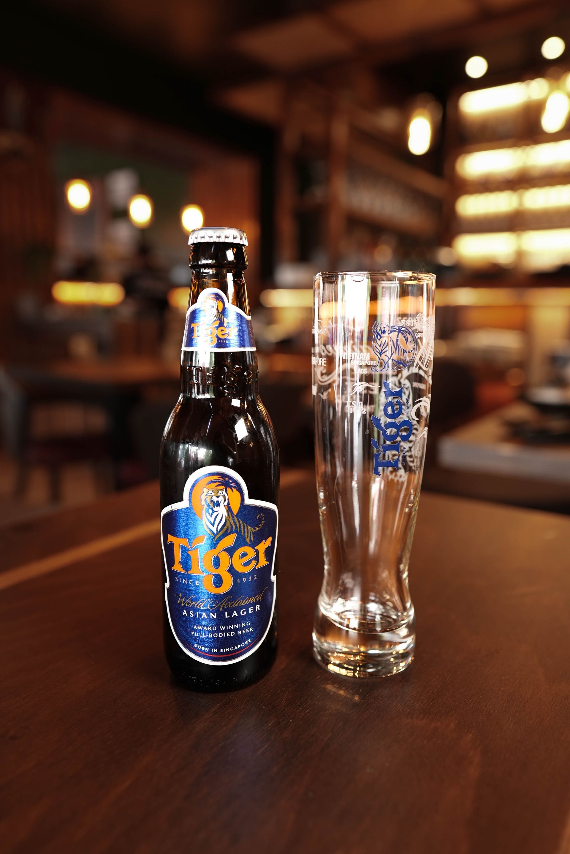Eine Tiger-Bierflasche und ein leeres Glas auf einem Bartisch, im Hintergrund warmes Licht.