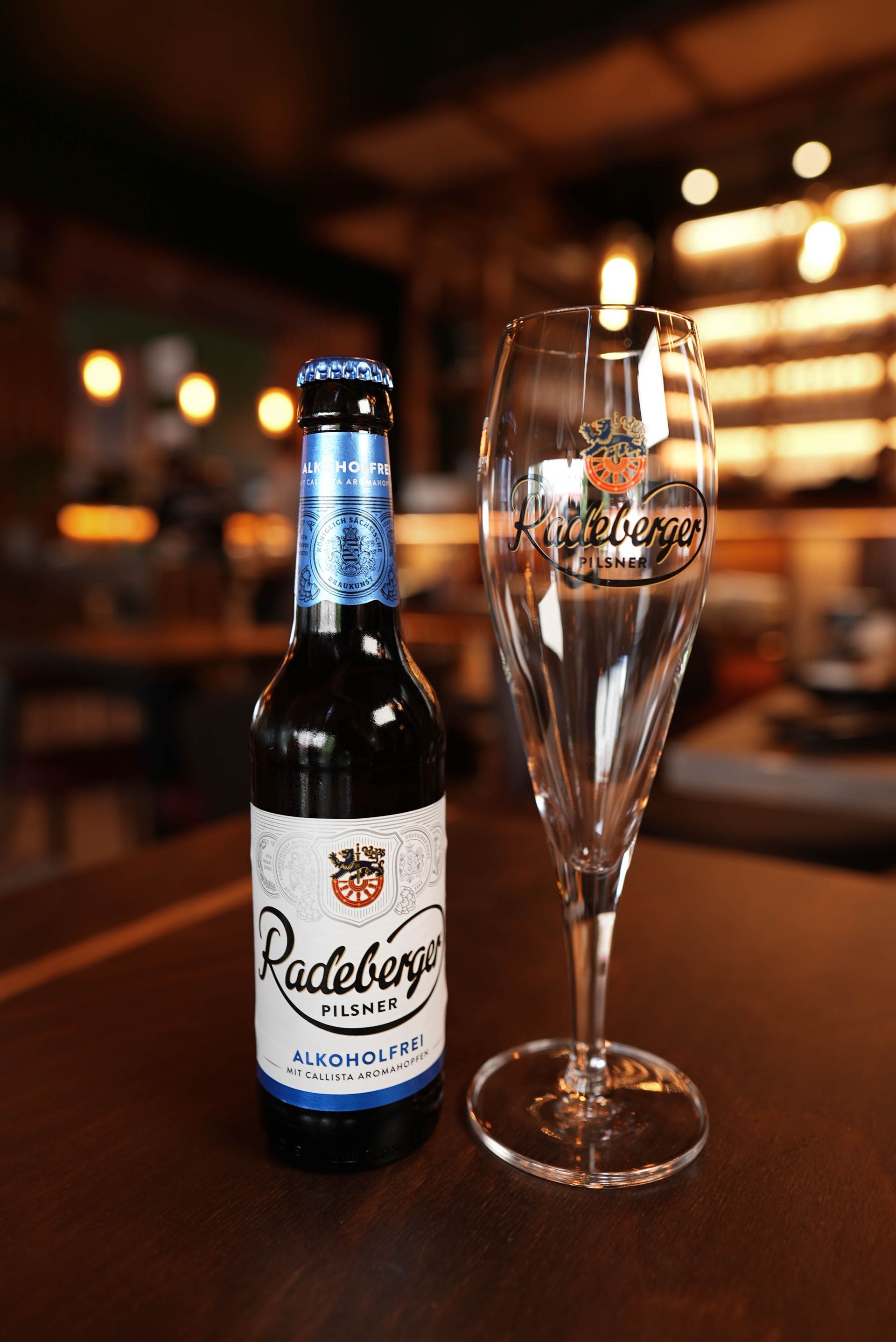 Eine Flasche Radlerbier neben einem leeren Sektglas auf einem warm beleuchteten Bartisch.