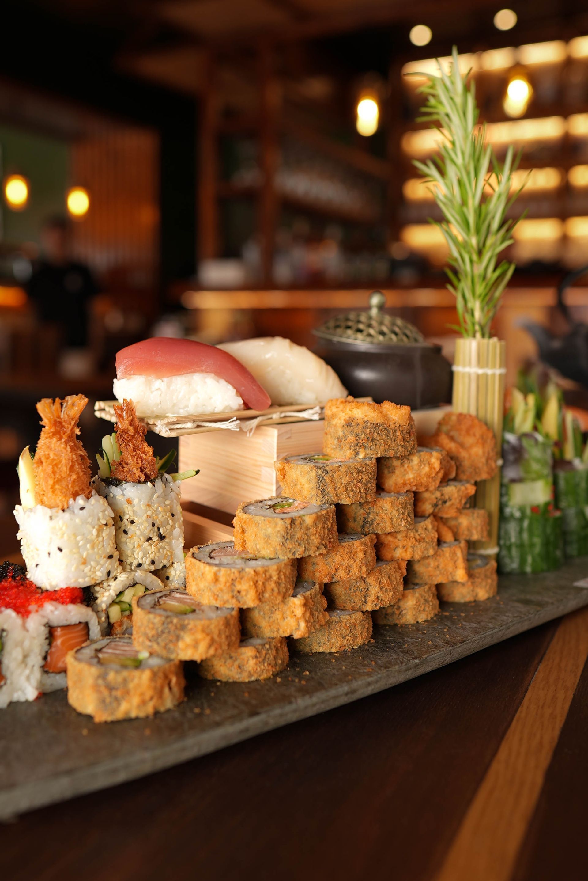 Verschiedene Sushi-Rollen und Nigiri auf einer Schieferplatte in einem Restaurantambiente, im Hintergrund warmes, verschwommenes Licht.
