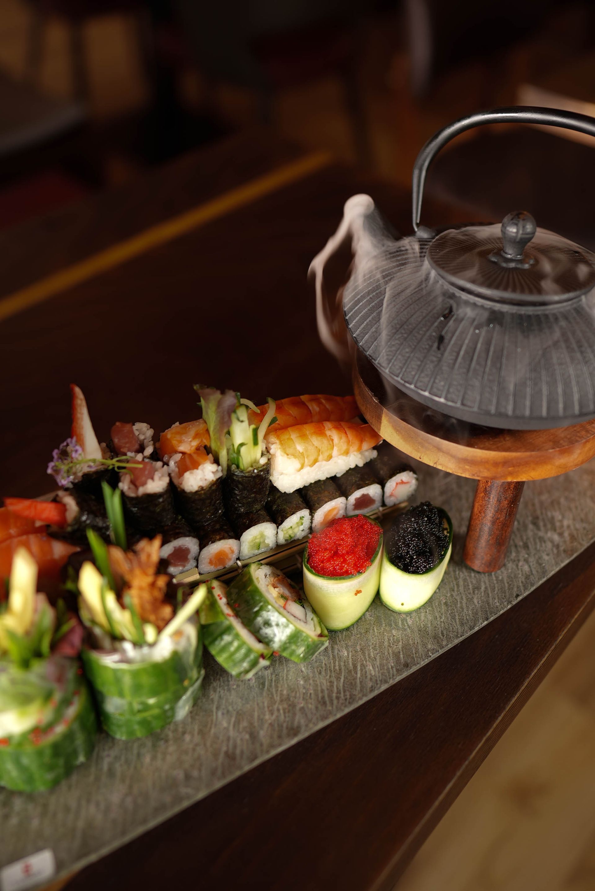 Sushi-Platte mit verschiedenen Rollen, Erdbeeren und einer Glasteekanne auf einem dunklen Serviertablett
