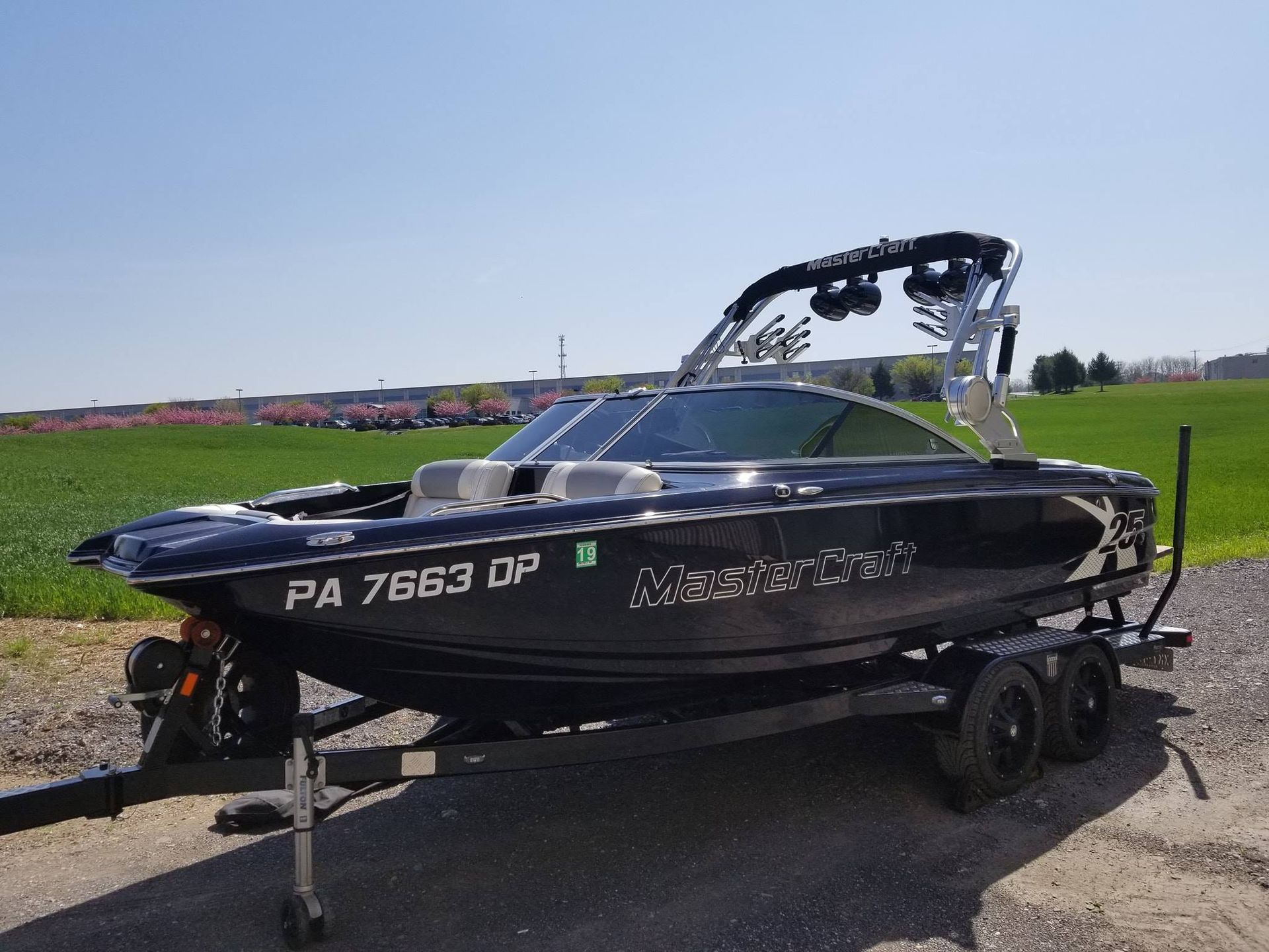 A black MasterCraft wakeboard boat on a trailer parked in a grassy field under a clear blue sky.