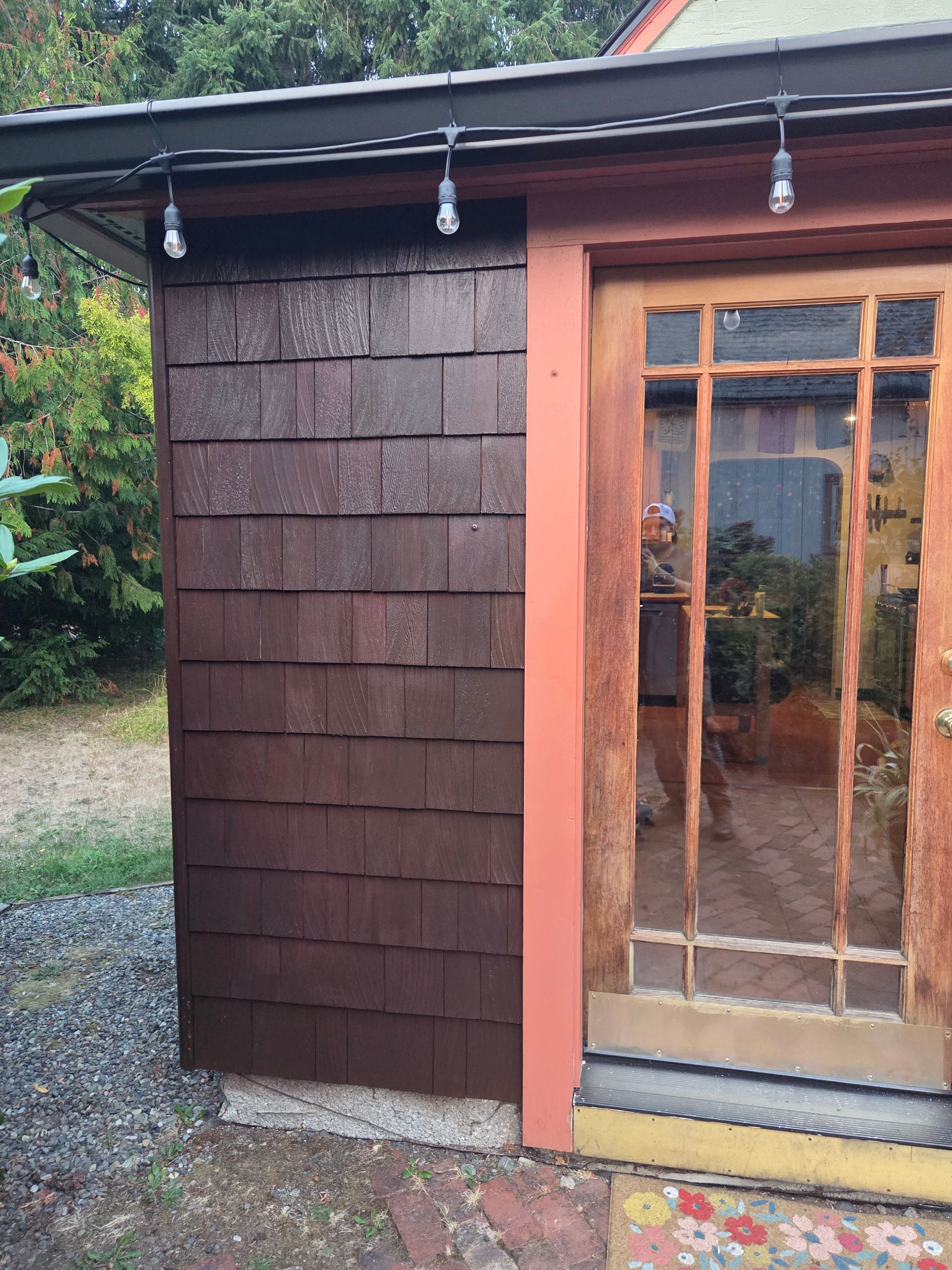 A wooden door with a glass pane sits next to a dark brown shingled wall under a string of outdoor lights.