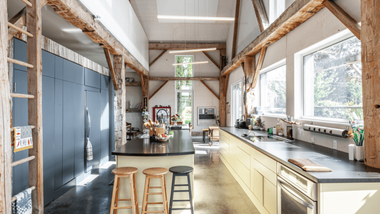 A high-ceilinged rustic kitchen with exposed wooden beams, a central island with stools, and cream-colored cabinetry.