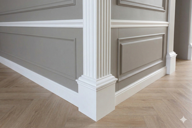 A corner wall with gray wainscoting, white decorative trim, and a white fluted pillar over a herringbone wood floor.