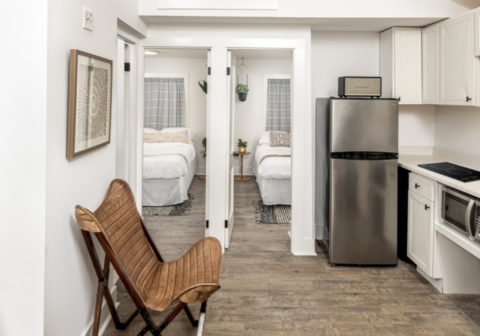 Small kitchen with stainless steel fridge, two bedrooms visible. Wicker chair in the foreground.