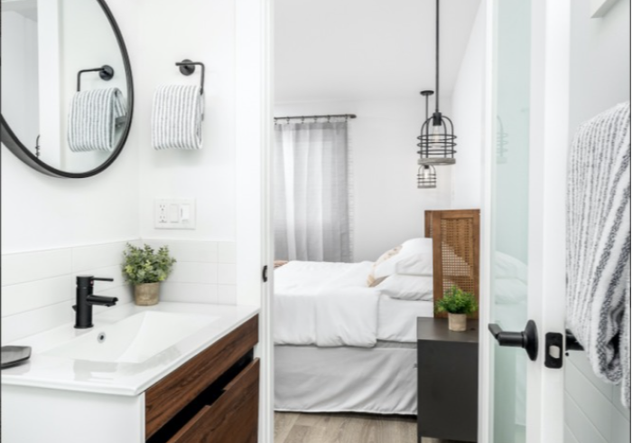 Bathroom doorway revealing a bedroom. White walls, wood vanity, round mirror, black hardware, bed, and sheer curtains.