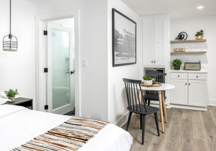 Small studio apartment with bed, bathroom, kitchenette, and dining area in white and wood tones.