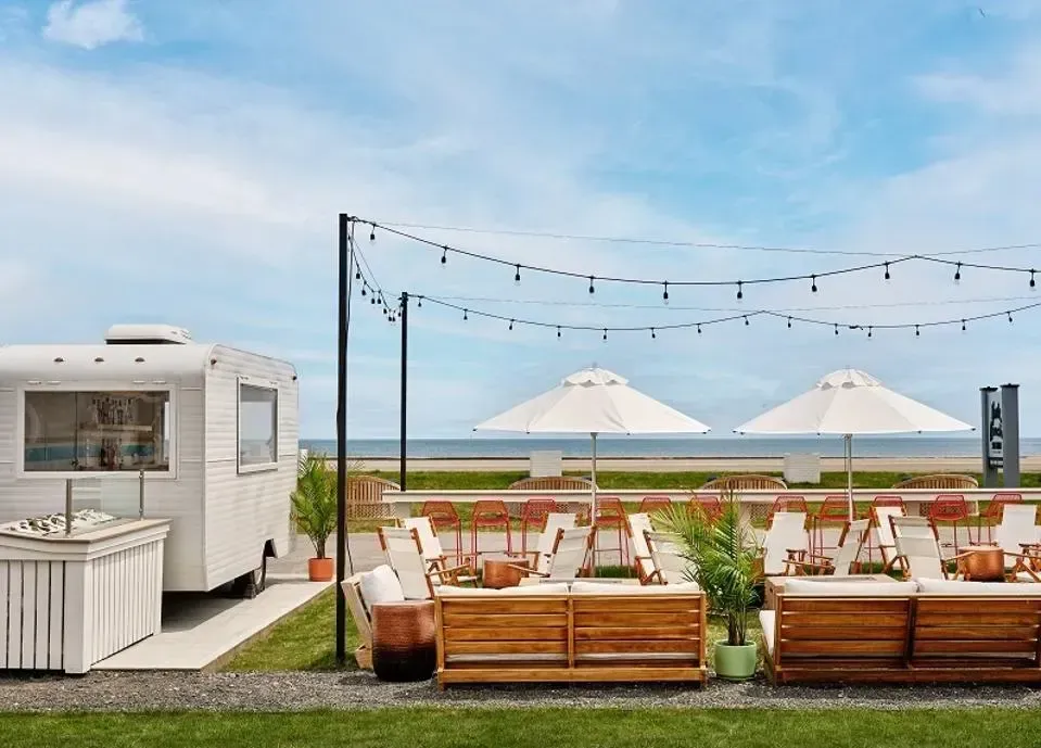 White food truck and outdoor seating with umbrellas on a grassy area, ocean in background.