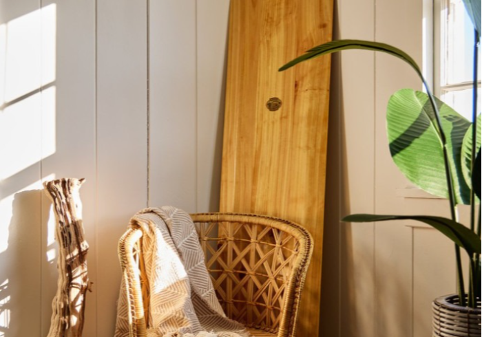 Wicker chair with blanket, leaning wooden board, and a potted plant by a window with sunlight.
