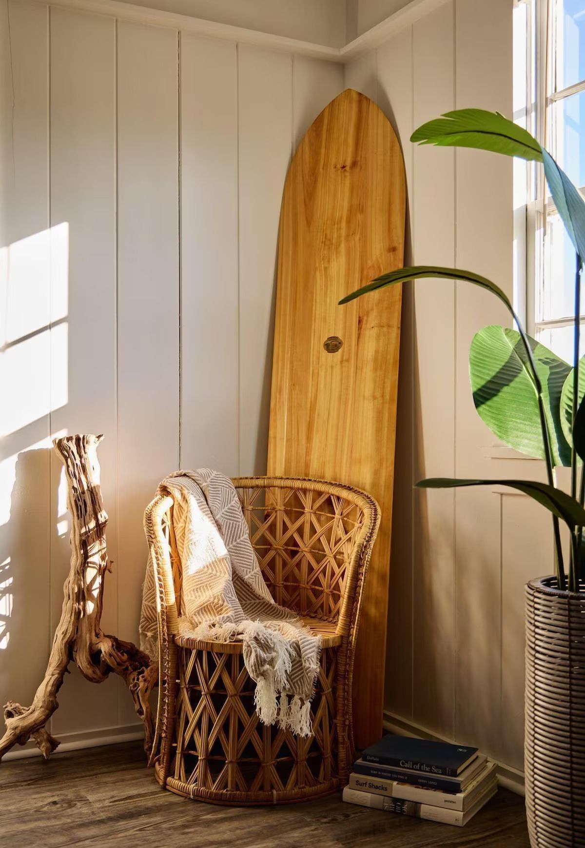 Wicker chair with blanket, wooden surfboard, driftwood, and potted plant in a corner of a sunlit room.