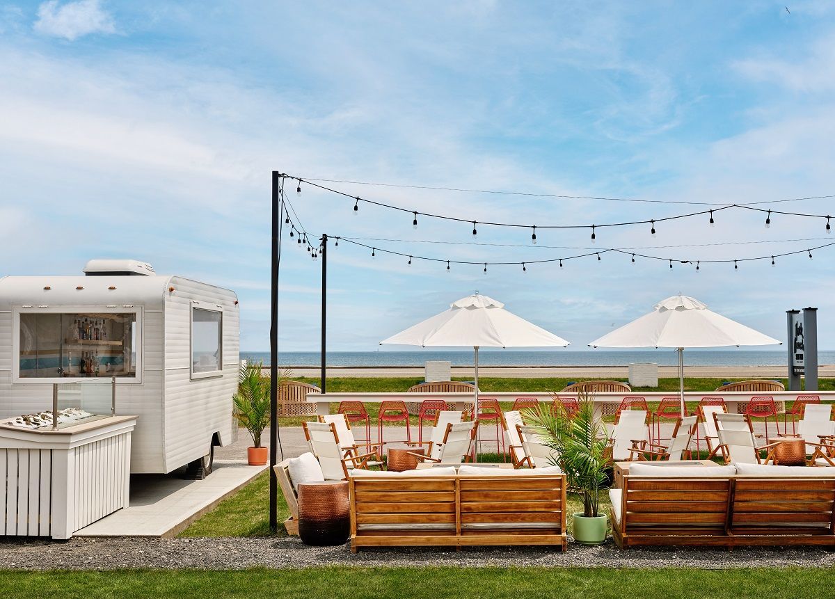 Outdoor event with trailer bar, tables, umbrellas, string lights, and ocean view.