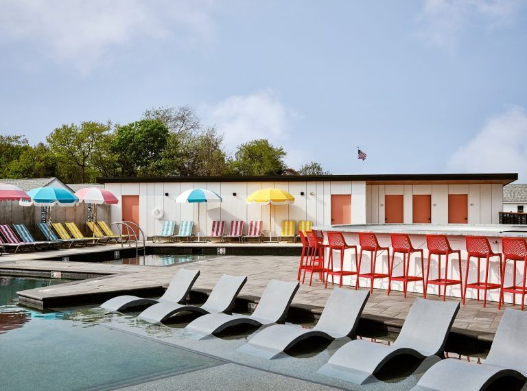 Poolside bar area with red stools, white loungers, and colorful umbrellas under a partly cloudy sky.