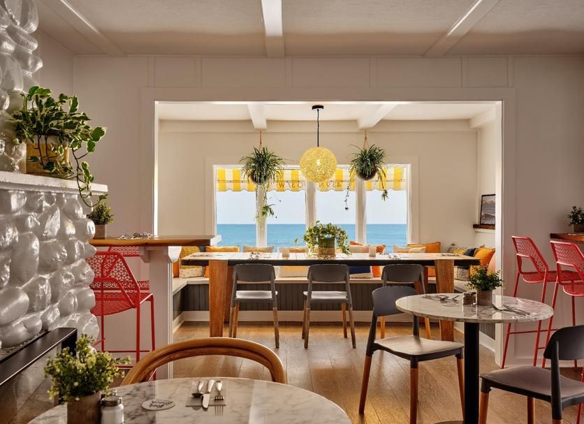 Restaurant interior with dining tables, chairs, and a long table overlooking the ocean through large windows.