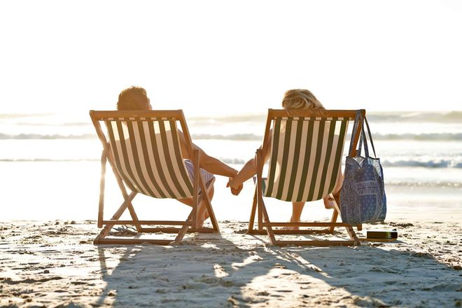 Two people sitting in wooden striped beach chairs, holding hands and facing the ocean on a sandy beach.