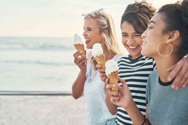 Three people laughing while eating ice cream cones on a beach boardwalk.