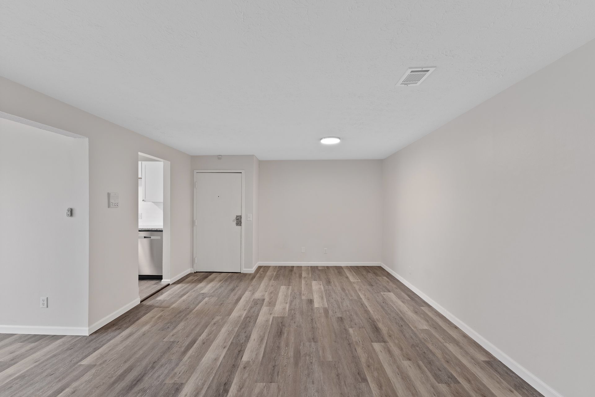 Open apartment living area with wood-style flooring and neutral walls leading to the kitchen.