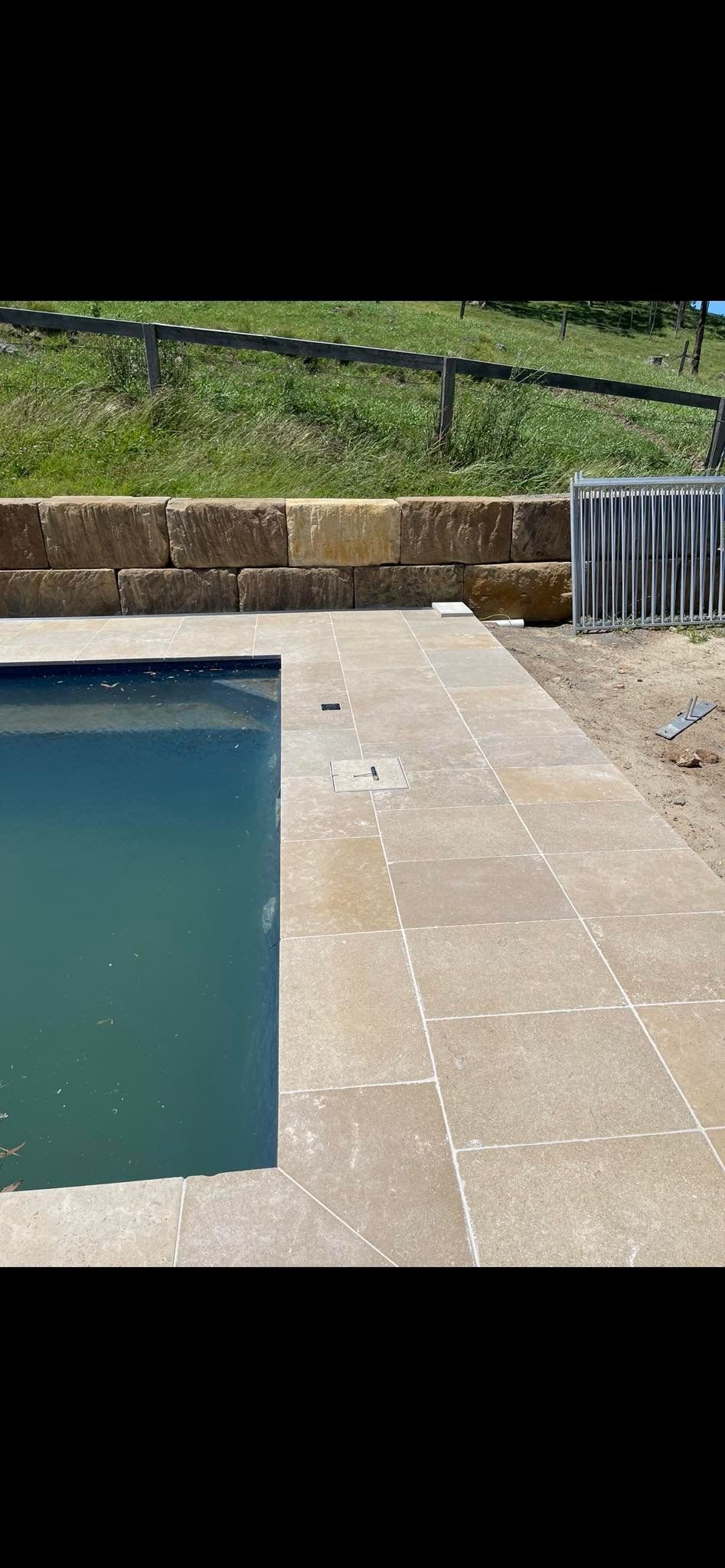 A swimming pool with tan stone paving and a retaining wall, with a grassy area in the background. — East Coast Paving in Seaham, NSW