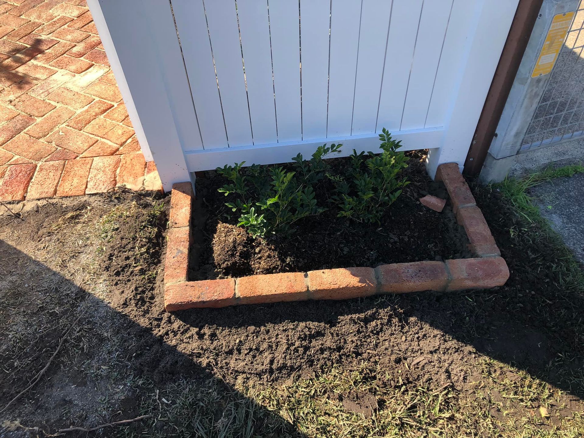 Brick-lined garden bed with small green plants against a white fence on brick pavers. — East Coast Paving in Seaham, NSW