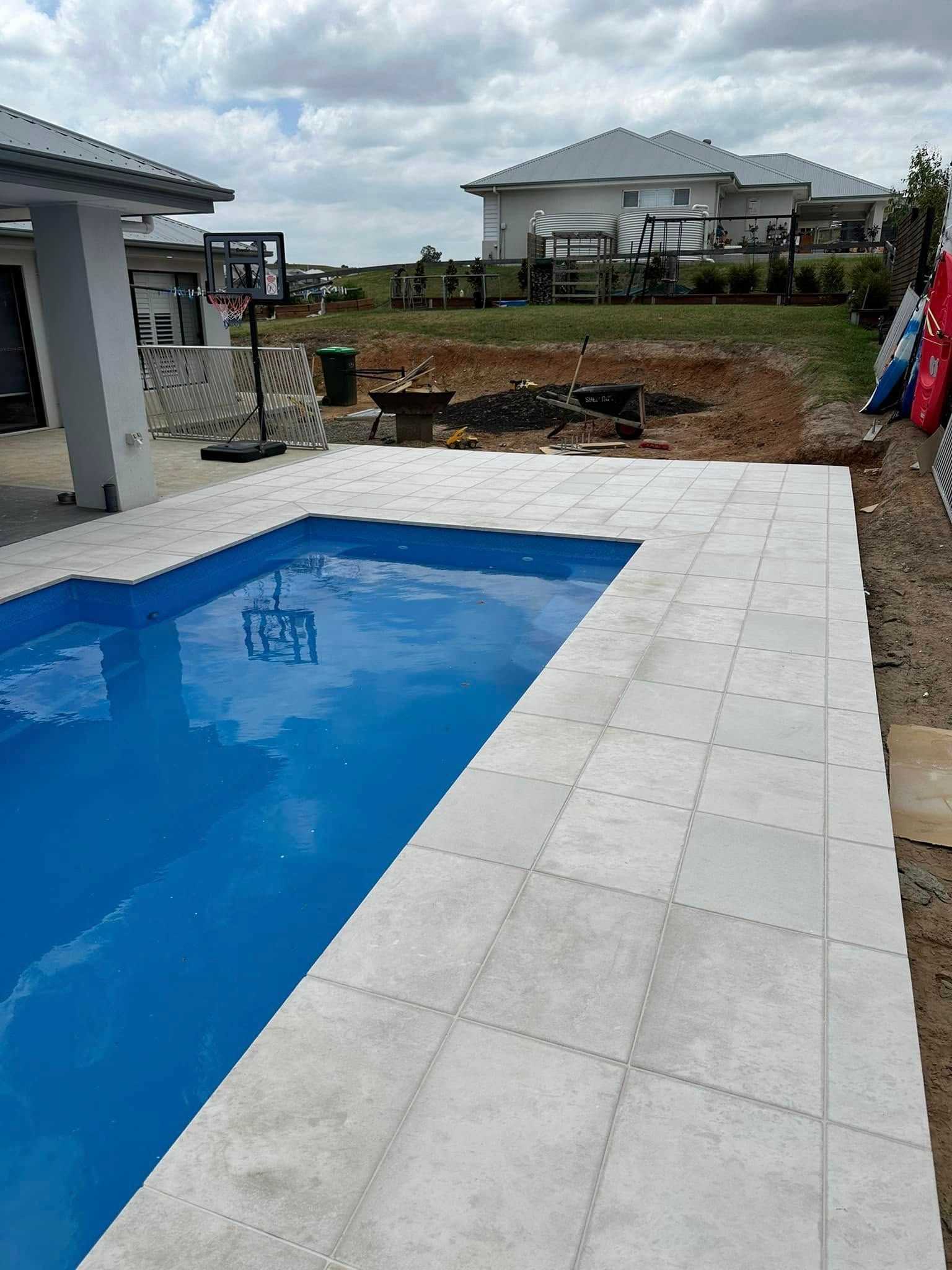 Pool with blue water and light gray patio tiles. Houses and sky in background. — East Coast Paving in Seaham, NSW