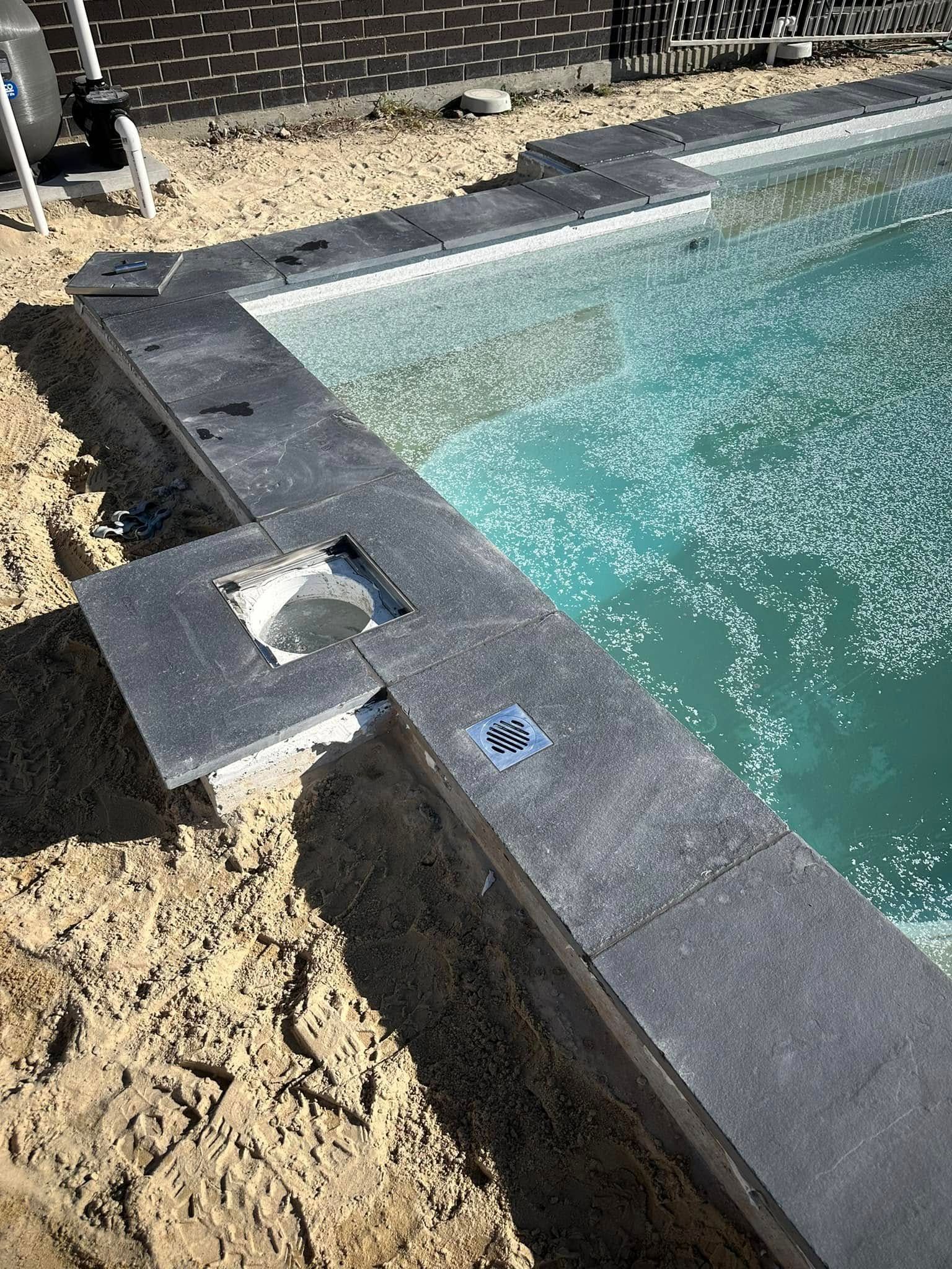 Pool under construction; dark gray border with water; sand in foreground, fence in background. — East Coast Paving in Seaham, NSW