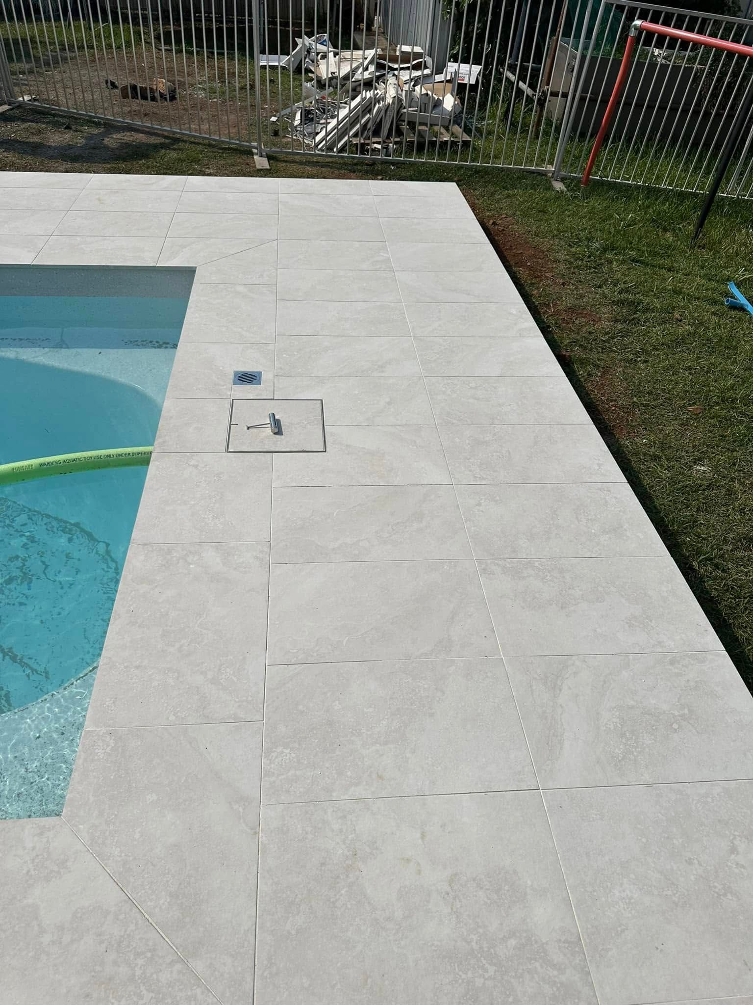 Poolside with light gray tiles and grass. A pool is on the left, and construction materials are in the background. — East Coast Paving in Seaham, NSW