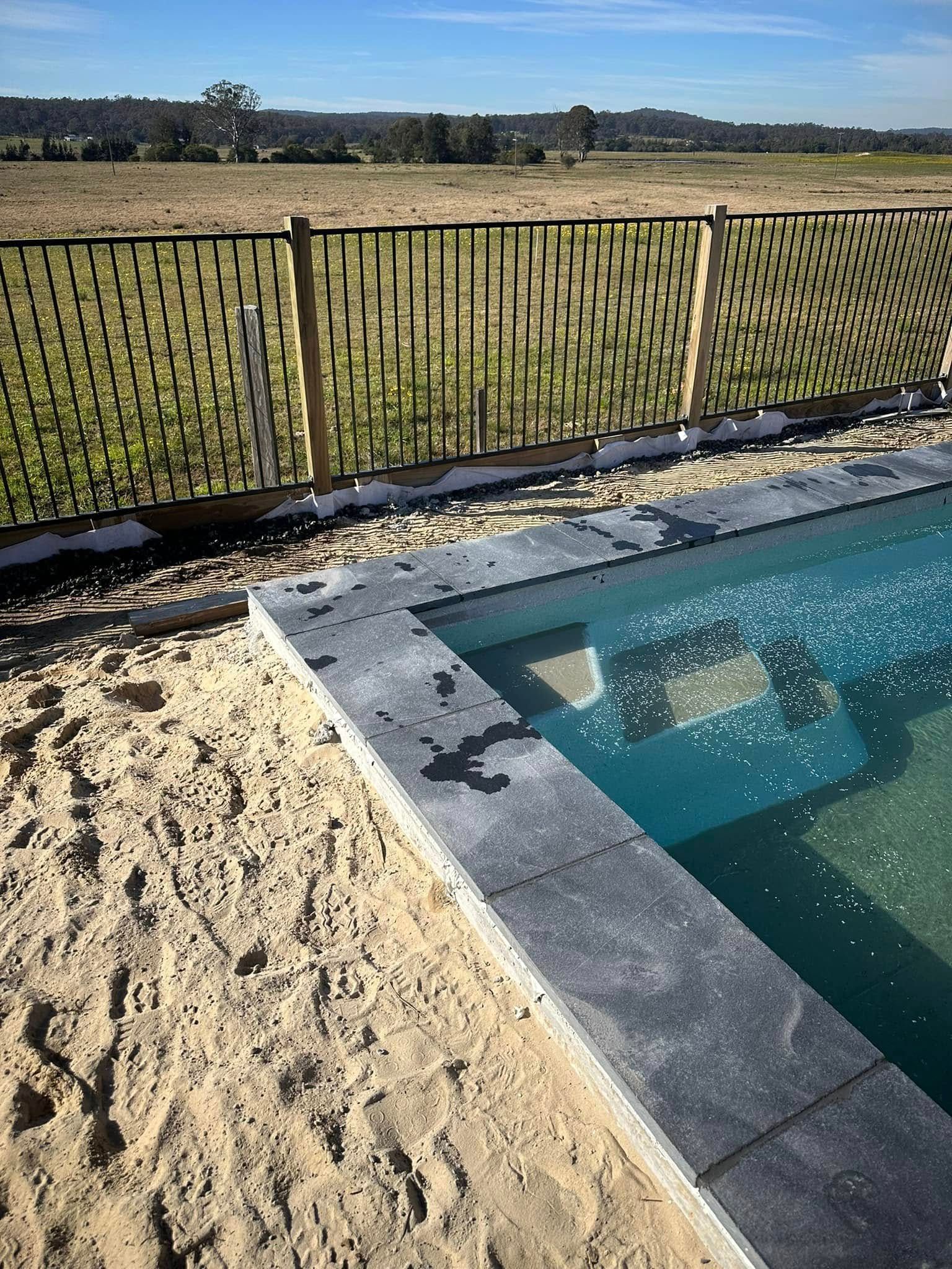 Pool with stone coping, sand, and woven black fence in a rural setting under a blue sky. — East Coast Paving in Seaham, NSW