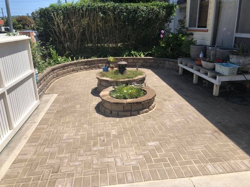 Brick patio with two tiered planters, a white fence, and greenery in a sunny setting. — East Coast Paving in Seaham, NSW