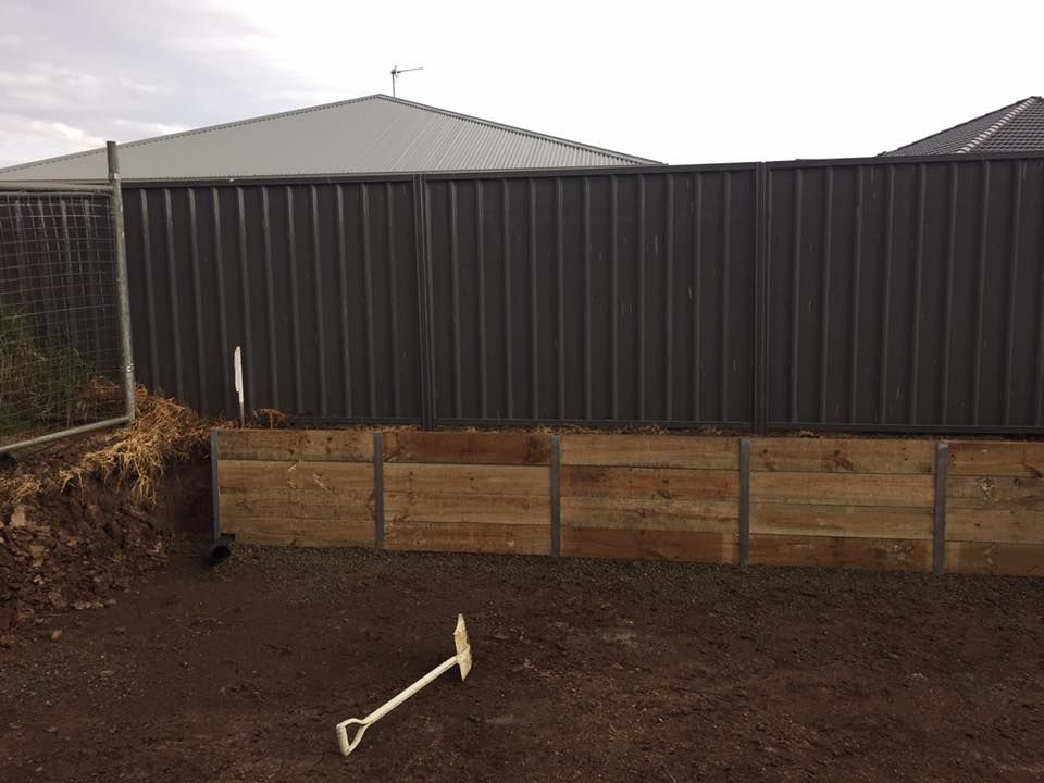 Wooden retaining wall in front of a corrugated metal fence, shovel in foreground, cloudy sky. — East Coast Paving in Seaham, NSW