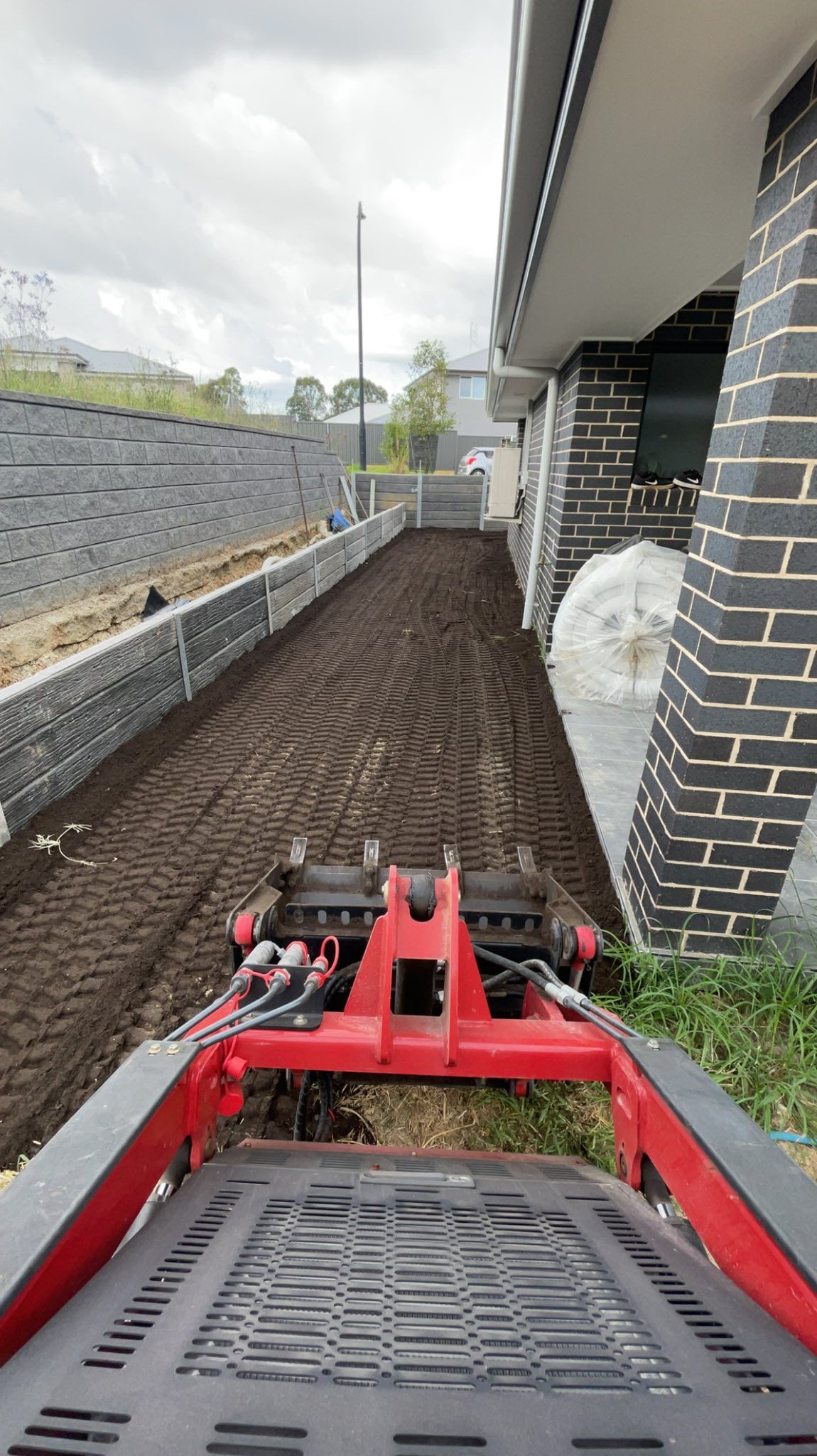 A red compact tractor tills freshly turned soil beside a modern building and retaining wall under a cloudy sky. — East Coast Paving in Seaham, NSW