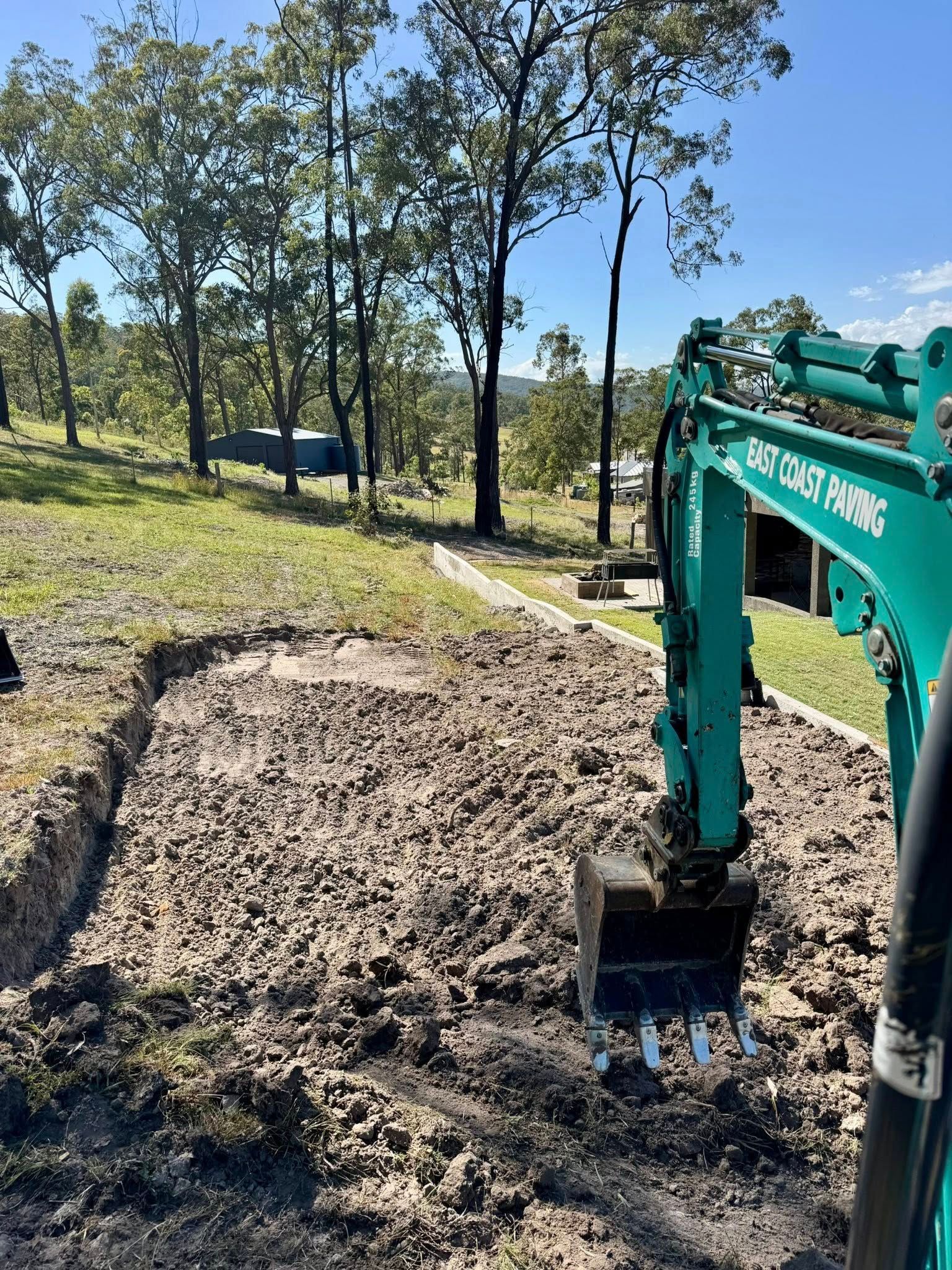 An excavator digging into dirt in a grassy area with trees in the background under a blue sky. — East Coast Paving in Seaham, NSW