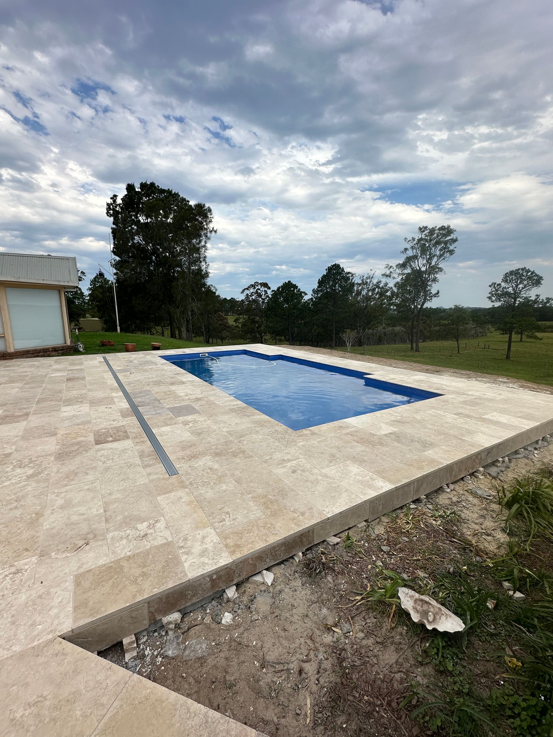 Swimming pool with blue water surrounded by beige tiles, under a cloudy sky.
