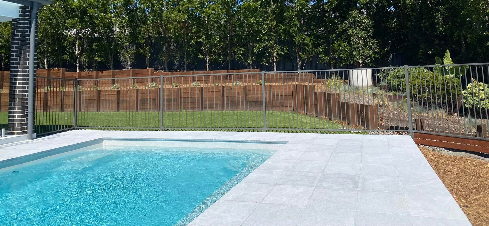 Swimming pool with patio and a grassy area, backed by a wooden fence and trees, under a blue sky.