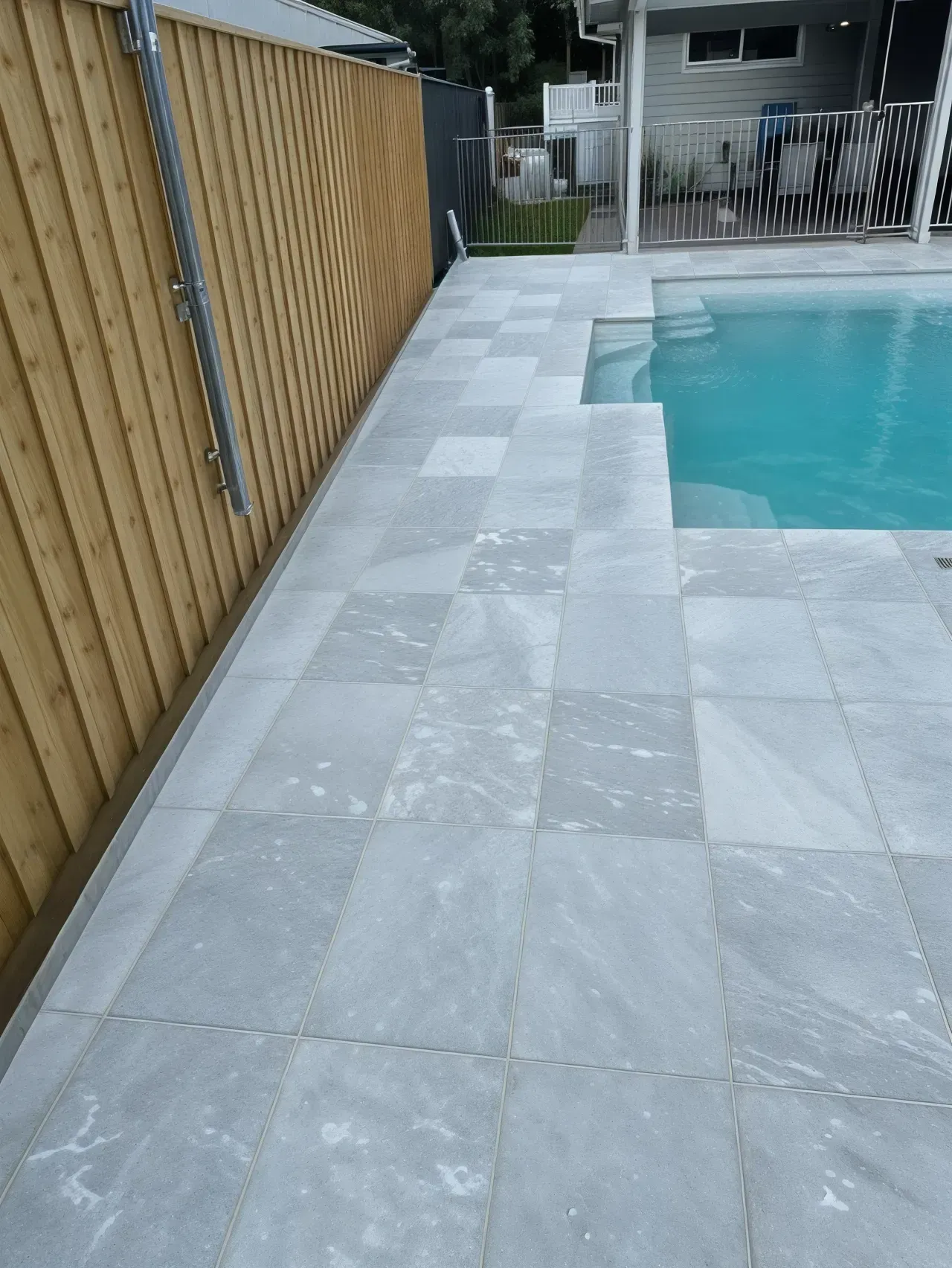Poolside with light gray tiled paving next to a wooden fence and pool. — East Coast Paving in Seaham, NSW