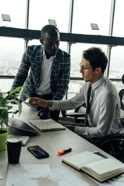 Two coworkers discuss a laptop at a desk in a bright office, with notebooks and a coffee cup nearby.