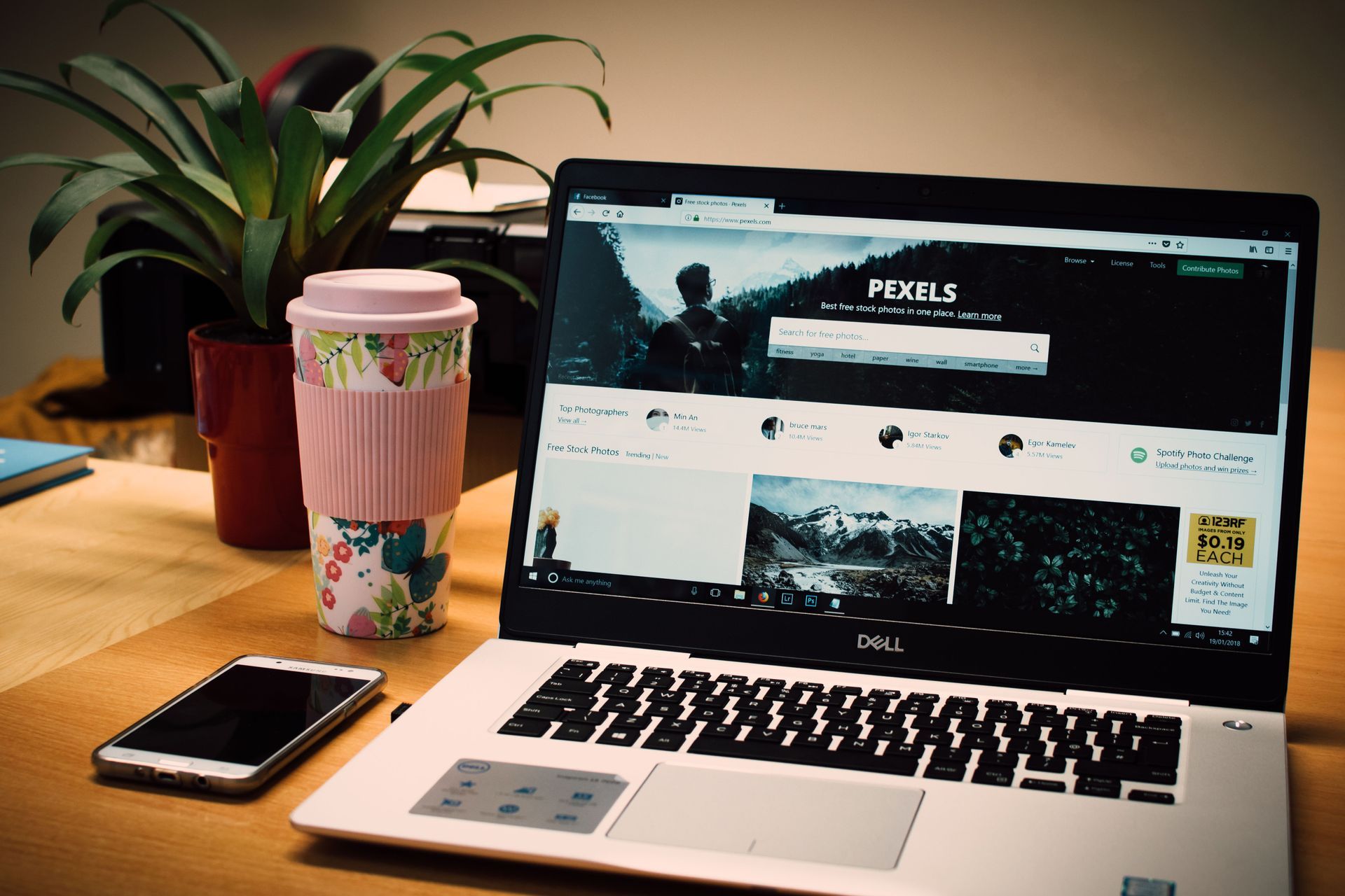 Laptop on desk with a webpage open, beside a phone, floral cup, and potted plant