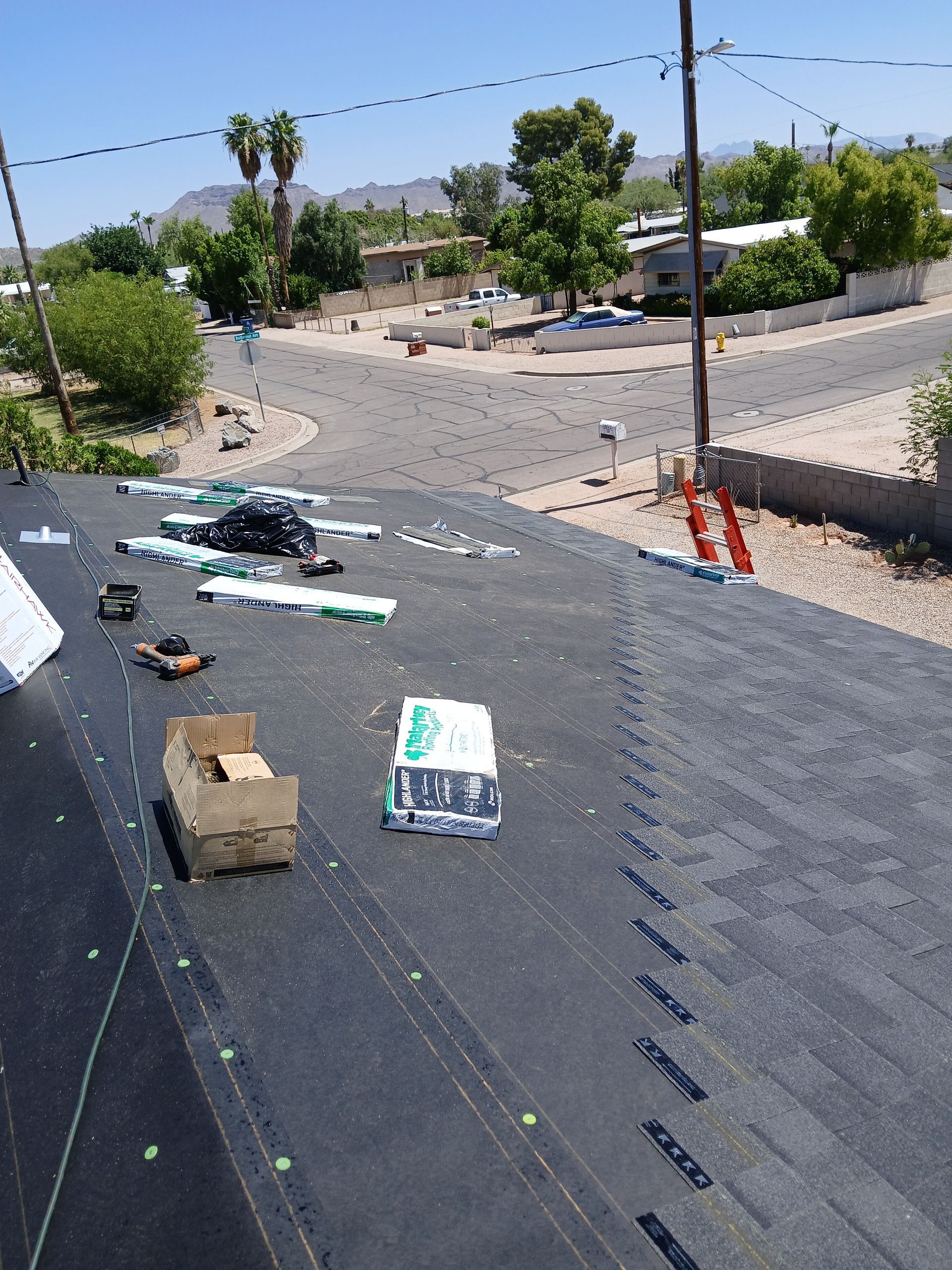 Rooftop with construction materials, including shingles, tools, and cardboard boxes. Suburban neighborhood in background.