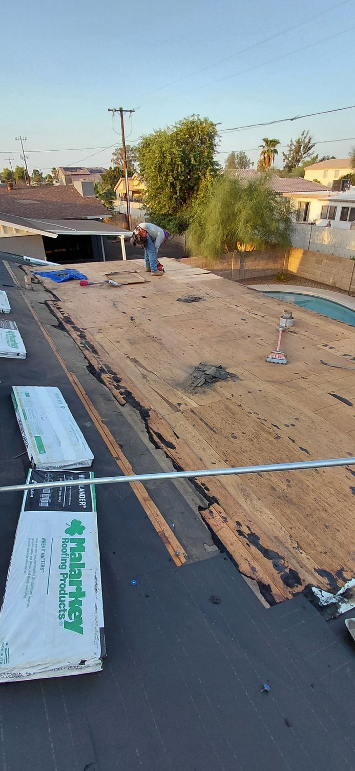 A worker on a rooftop removes old roofing material. Supplies and homes are in the background.