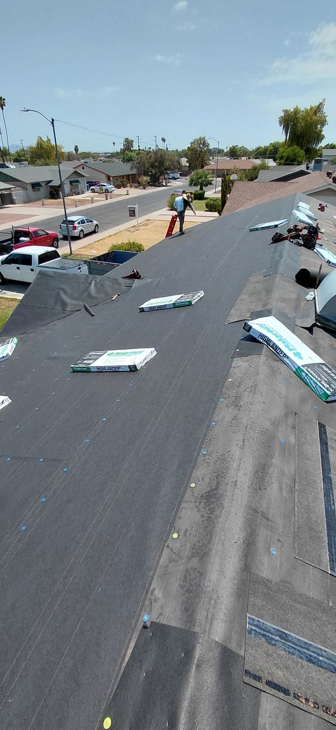 Workers on a rooftop installing shingles with a neighborhood street in the background on a sunny day.