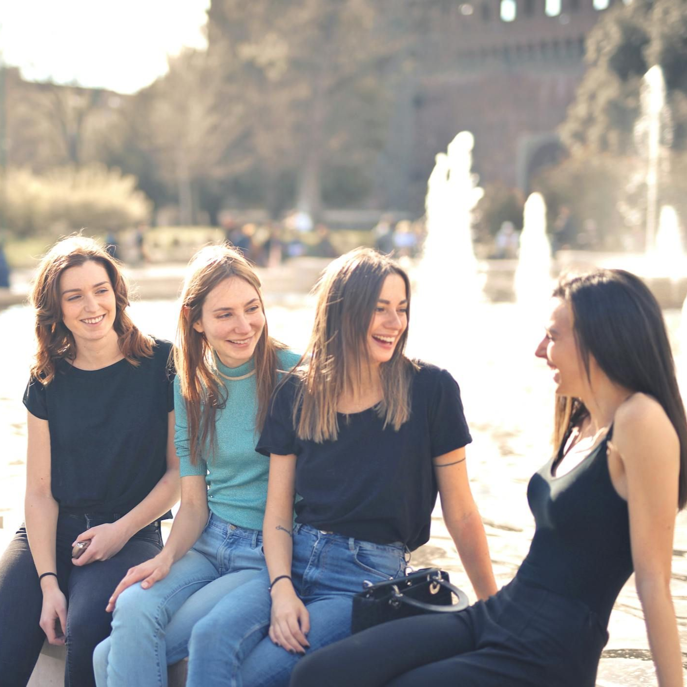 Four friends sit and laugh together by a fountain in a park, enjoying a sunny day.