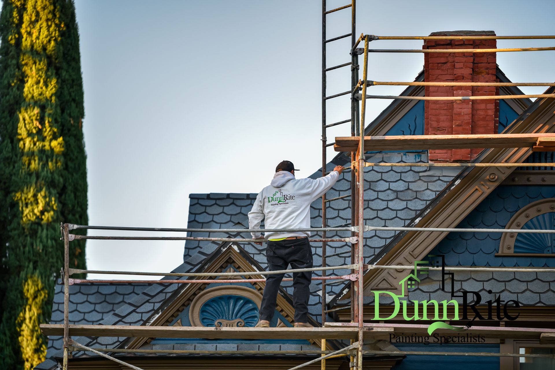 Man in Redlands, CA on scaffolding painting house trim, blue and gray house with red brick chimney.