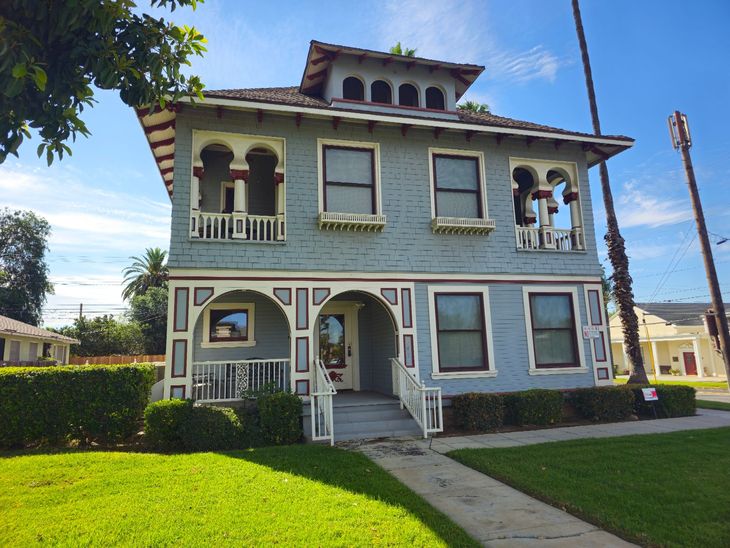 We painted A two-story blue house with ornate arched balconies, a hip roof, and a front porch, surrounded by green lawns and trees in Redlands.