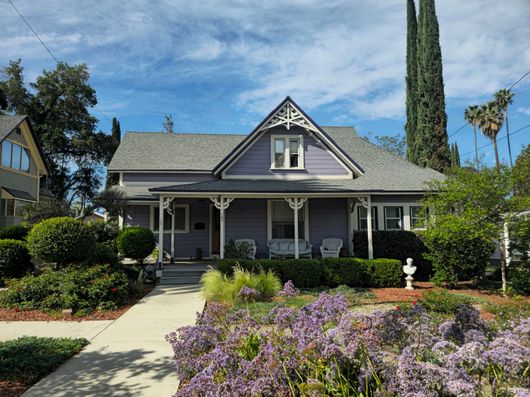 A light purple Victorian-style house with a covered front porch, set behind a flower garden and a concrete walkway in Redlands, CA.