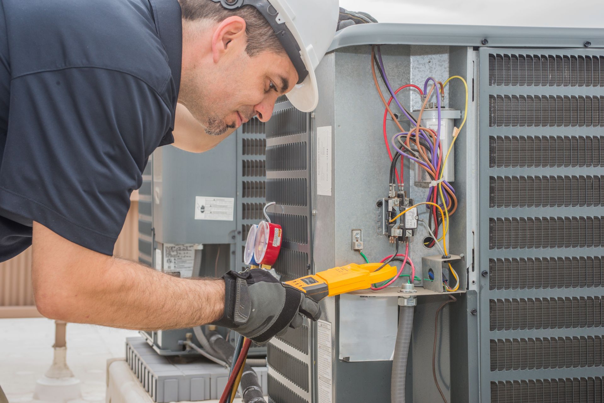 HVAC technician in a hard hat, examining AC unit wiring with a multimeter. Outdoor setting.