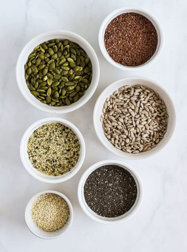 Cuencos de diversas semillas sobre una superficie blanca: calabaza, lino, girasol, cáñamo, sésamo y chía.