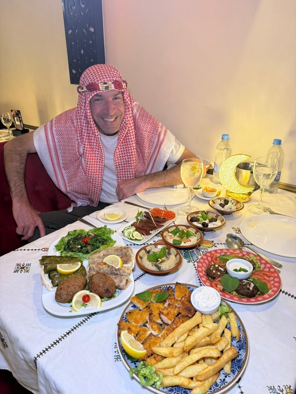 Un hombre sentado a una mesa con una gran variedad de comida y bebida, sonriendo en el interior de una vivienda.