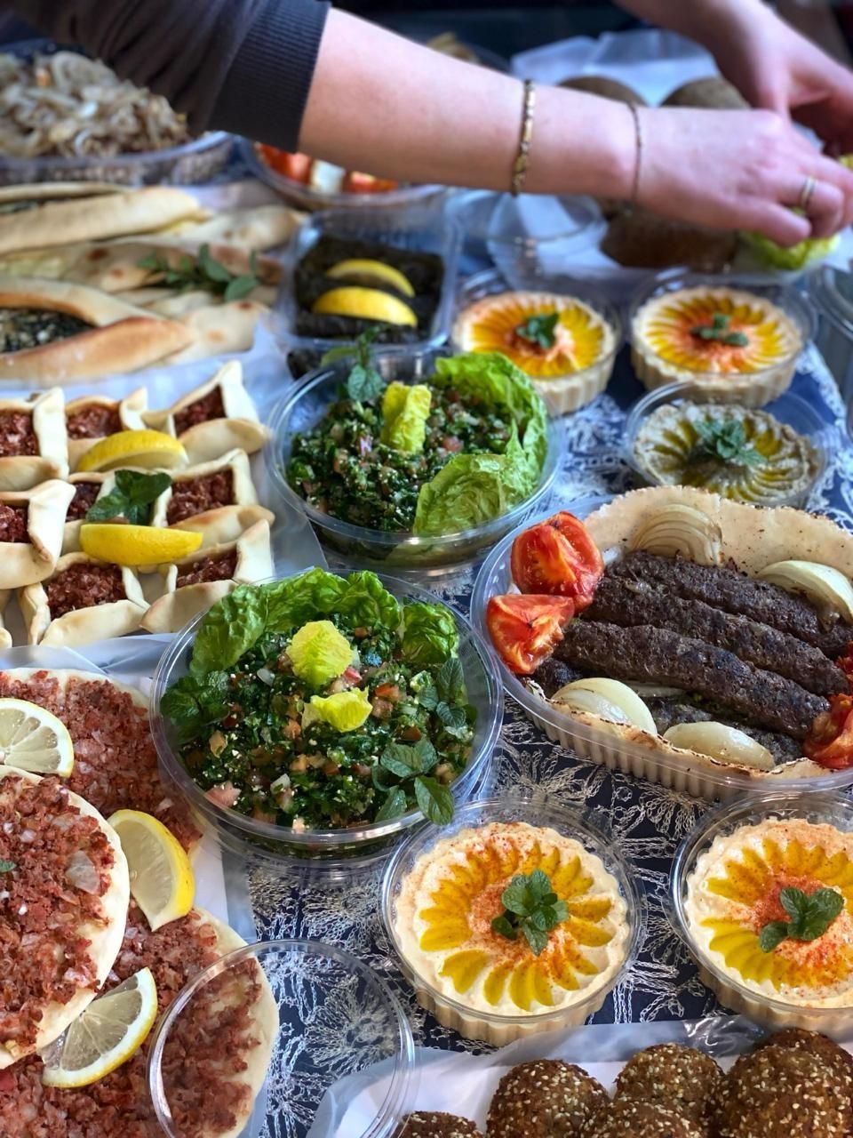 Manos colocando un bufé de coloridas ensaladas, pasteles y platos rellenos sobre una mesa.